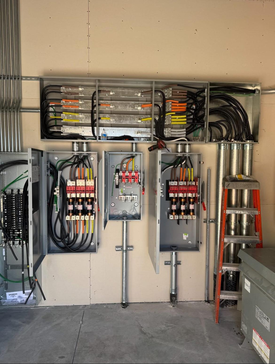 Commercial service wall with an overhead splitter trough, three fused disconnect switches, a Siemens panelboard, and a dry-type transformer