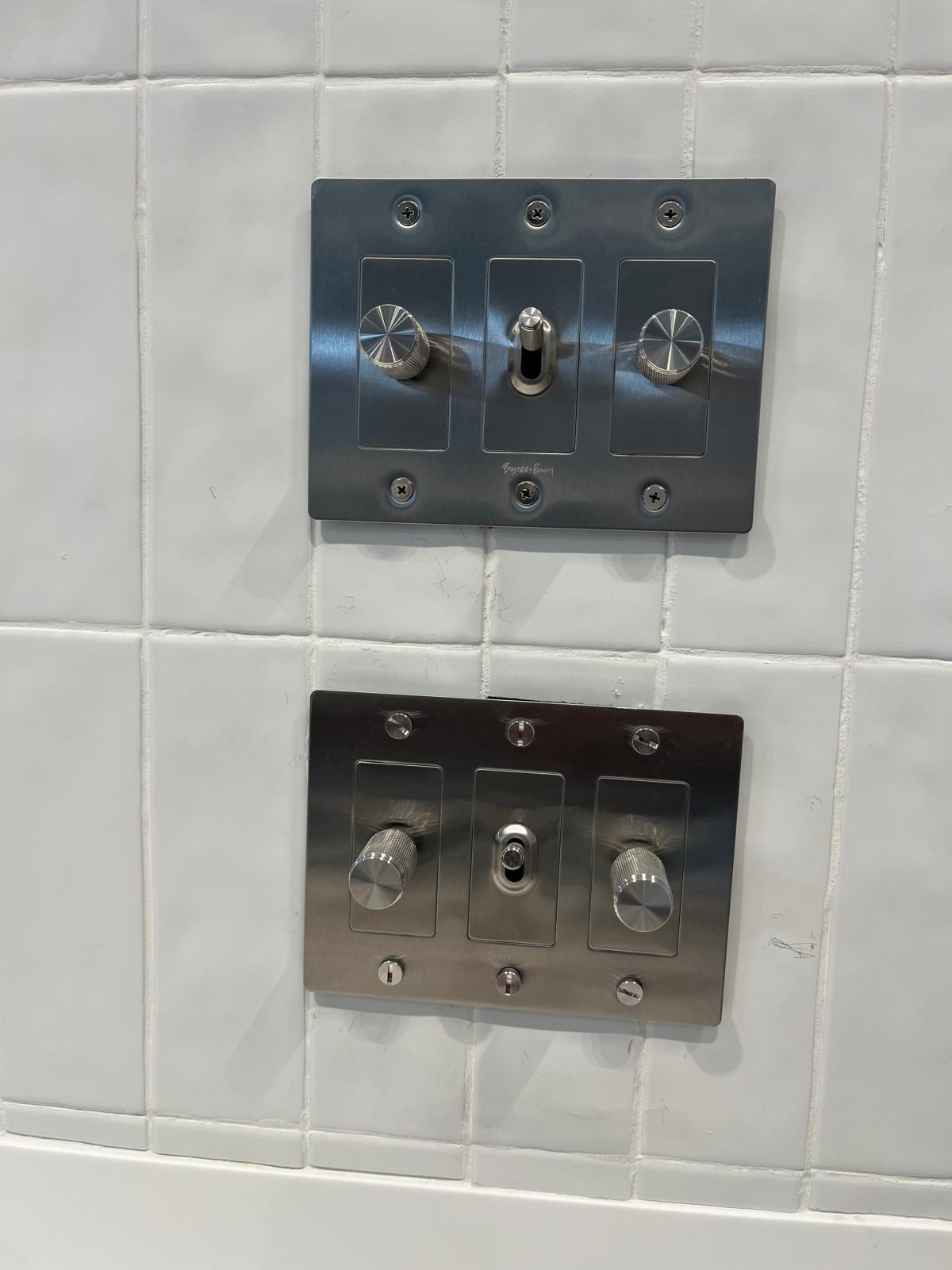 Two brushed steel three-gang switch plates with knurled dimmer knobs and toggle switches, mounted square on white tile