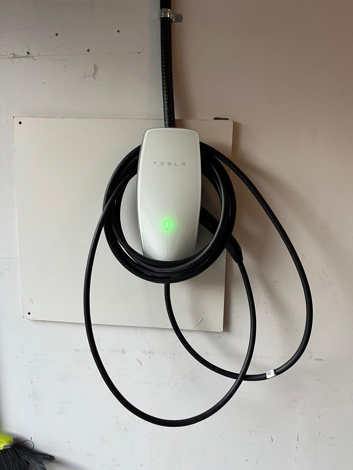 Wall-mounted Tesla Wall Connector energized with a green status light, fed by flexible conduit, cable coiled on the mount