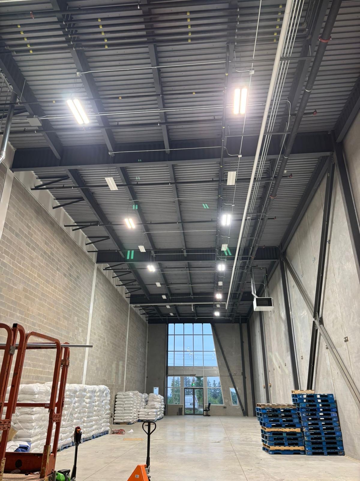 Warehouse interior with rows of linear LED light fixtures and conduit run across open steel joists at full ceiling height